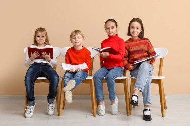 Little children reading books while sitting on chairs near beige wall