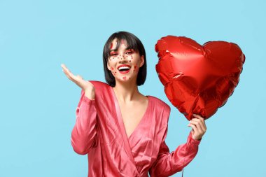 Beautiful young woman with creative makeup for Valentines Day and heart-shaped balloon on blue background