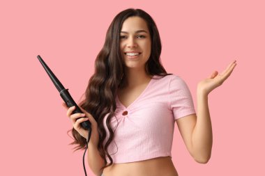 Beautiful young woman with curling iron on pink background