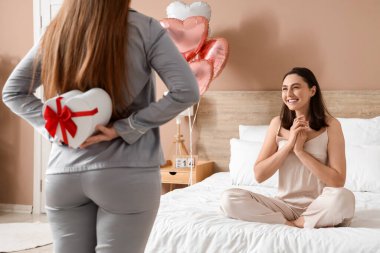 Young lesbian woman and her beloved hiding gift behind back in bedroom on Valentine's Day