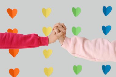 Young lesbian couple holding hands against rainbow hearts on light background, closeup. Valentine's Day celebration