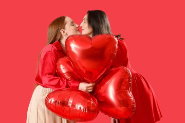 Young lesbian couple with heart-shaped balloons on red background. Valentine's Day celebration