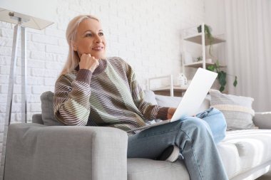 Evde kanepede dizüstü bilgisayarla çalışan olgun bir kadın programcı.