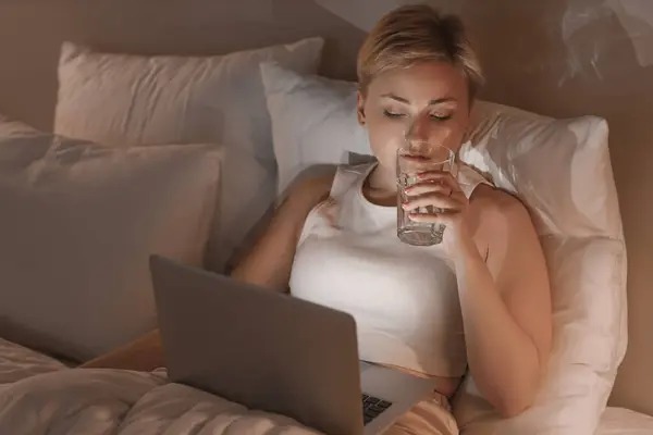 Young woman with laptop drinking water in bed at night