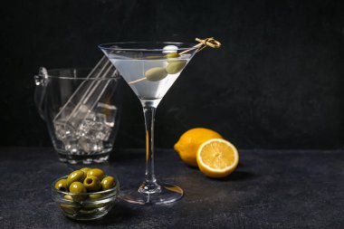 Bir bardak lezzetli martini, limon, buz küpleri ve koyu arkaplanda yeşil zeytinler.