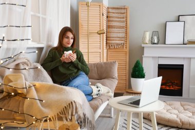 Elinde bir fincan kakao olan genç bir kadın evde video izliyor.