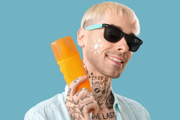Tattooed man with sunscreen cream on light blue background