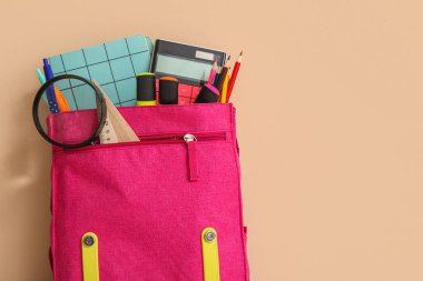 Pembe okul çantası, farklı kırtasiye malzemesi ve turuncu arka planda büyüteç.
