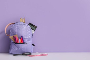 Okul çantasının içinde farklı kırtasiye malzemeleri ve mor duvarın yanındaki beyaz masada kulaklıklar var.