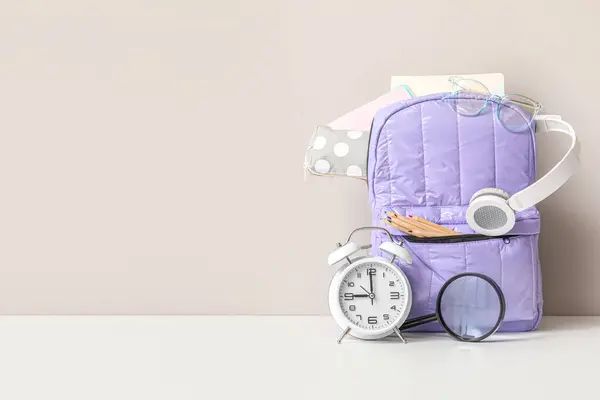 Purple school backpack with different stationery and alarm clock on white table near wall