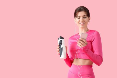 Kulaklıklı, su şişeli, pembe arka planda cep telefonu kullanan genç bir kadın.