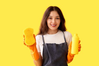 Sarı arka planda sünger ve deterjanı olan genç bir kadın.