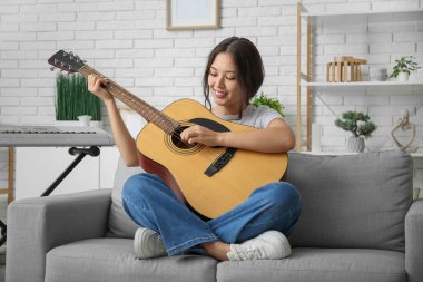 Evdeki kanepede otururken gitar çalan genç ve güzel bir kadın.