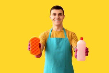 Sarı arka planda sünger ve deterjanı olan genç bir adam.