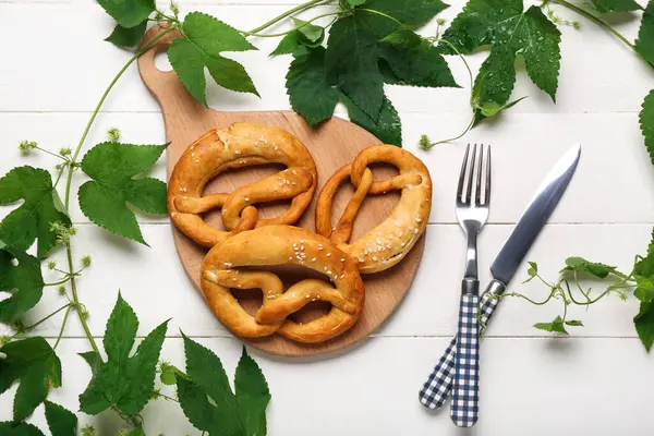 Board with soft pretzels and hop leaves on white wooden background