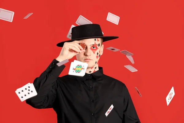 Young man with drawn suits on his face, poker chip and falling cards against red background
