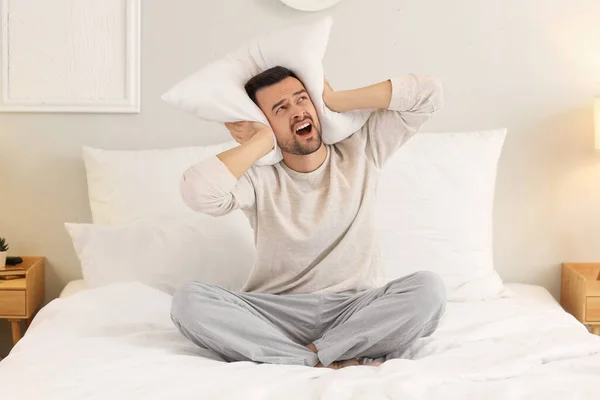 Young man with pillow suffering from loud neighbours in bedroom