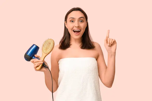 Beautiful young woman with hair dryer and brush pointing at something on pink  background