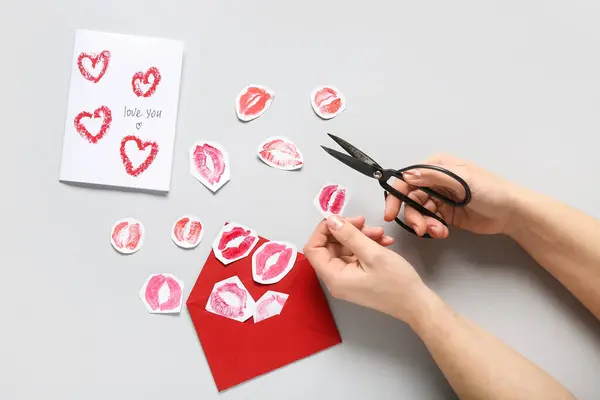 Female hands with paper lipstick kiss marks, scissors, card and envelope on grey background. Valentine\'s Day celebration