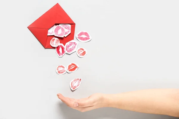 Female hand with red envelope and paper lipstick kiss marks on grey background, closeup. Valentine\'s Day celebration