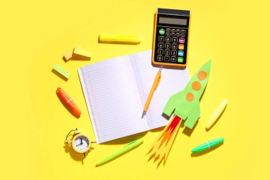 Hesap makineli kağıt roket, boş defter ve sarı zemin üzerinde farklı kırtasiye malzemesi