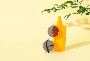 Güneş gözlüklü bir şişe güneş kremi ve soluk sarı arka planda ağaç dalı.