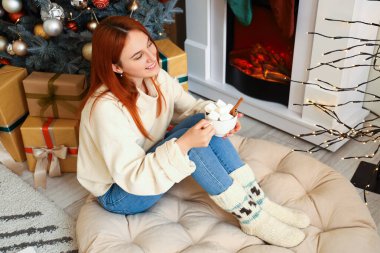 Genç bir kadın Noel arifesinde şöminenin yanında kakao içiyor.