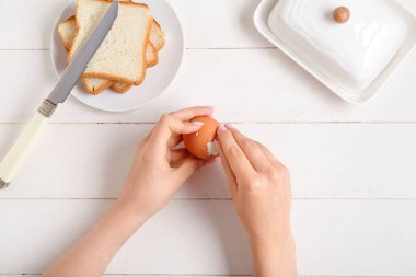 Beyaz ahşap arka planda haşlanmış tavuk yumurtası ve tabağı soyan kadın.