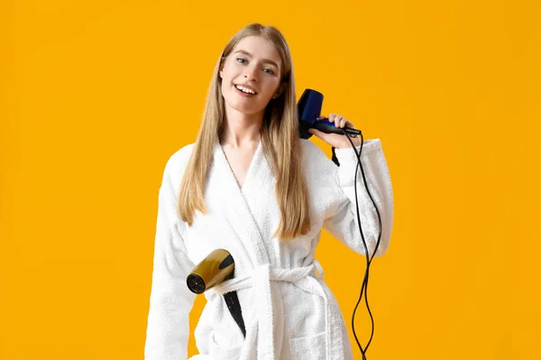 Young blonde woman with hair dryers on yellow background