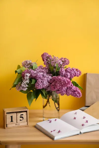 Vase with lilac flowers, calendar and books on table near yellow wall