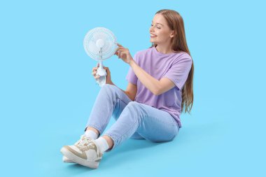Mavi arka planda oturan elektrikli fanı olan genç bir kadın.