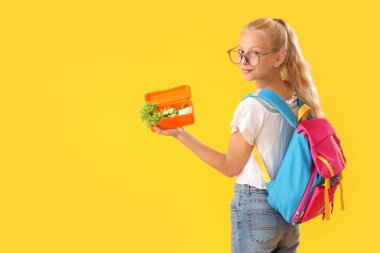 Sırt çantası ve sarı arka planda beslenme çantası olan gözlüklü mutlu bir kız.