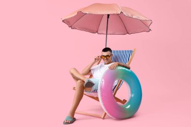 Şişirilebilir yüzüğü ve şemsiyesi olan mutlu genç adam pembe arka planda güverte sandalyesinde oturuyor.