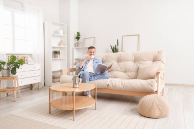 Evdeki kanepede bir fincan sıcak kahve okuyan olgun bir adam.