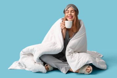 Yumuşak battaniyeli, uyku maskesiyle ve mavi arka planda oturan bir fincan kahveyle güzel, genç ve mutlu bir kadın.