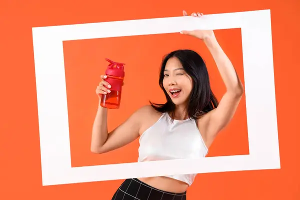 Young beautiful Asian woman with frame and bottle of water on orange background