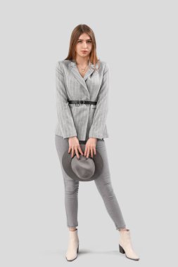 Gri arka planda şapkası olan, ceketli ve kot pantolonlu şık genç bir kadın.