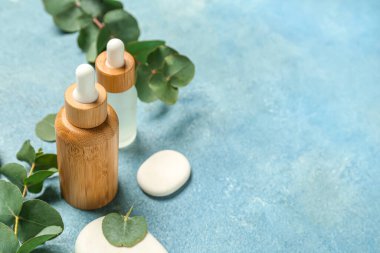 Kozmetik yağlı şişeler, okaliptüs dalı ve mavi desenli arka planda taşlar.