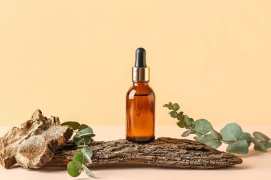 Bir şişe kozmetik yağı, okaliptüs dalları ve soluk sarı arka planda ağaç kabuğu.