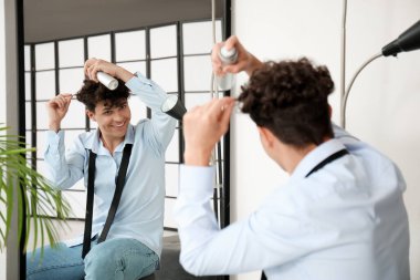 Yakışıklı genç adam aynanın yanındaki kıvırcık saçlarına saç spreyi sıkıyor.