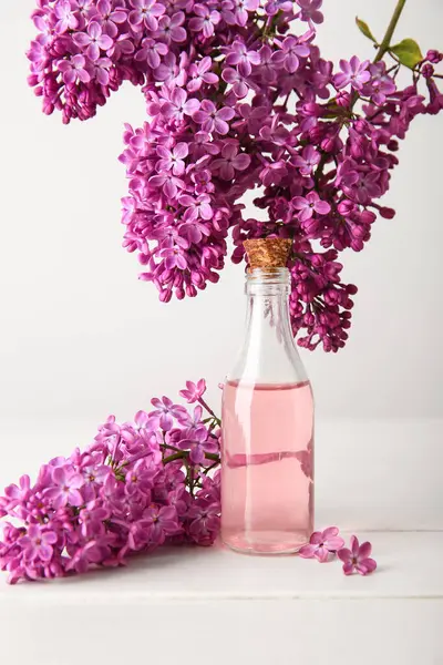 Bottle of perfume with beautiful lilac flowers on wooden table near white wall