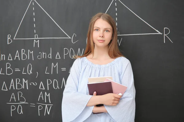 Young math teacher with books near blackboard in classroom