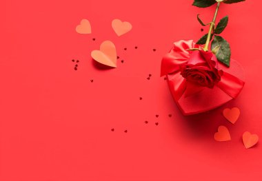 Güzel bir gül, kalp şeklinde bir hediye kutusu ve kırmızı arka planda kağıt kalpler. Sevgililer Günü kutlaması