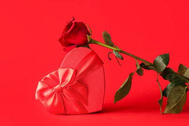 Güzel bir gül çiçeği ve kırmızı arka planda kalp şeklinde bir hediye kutusu. Sevgililer Günü kutlaması