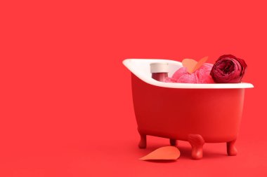 İçinde banyo malzemeleri, kağıt kalpler ve kırmızı arka planda gül olan mini bir küvet. Sevgililer Günü kutlaması