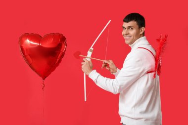 Aşk tanrısı gibi giyinmiş kırmızı arka planda balon ve yay olan genç bir adam. Sevgililer Günü kutlaması