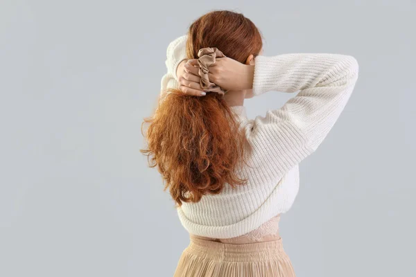Young redhead woman making ponytail with scrunchy on light background, back view