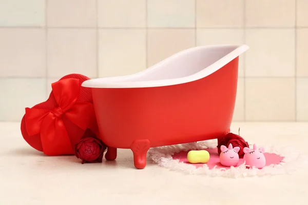 Mini bathtub with gift box, roses and rubber unicorns on white table near tile. Valentine's Day celebration