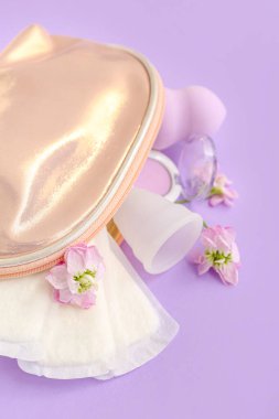 Leylak arka planında regl fincanı, ped ve makyaj ürünleri olan bir kadın makyaj çantası.