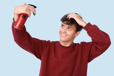 Kıvırcık saçlarına mavi arka planda saç spreyi sıkan yakışıklı bir genç.